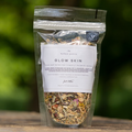 Herbal tea package labeled 'Glow Skin' by hulka pantry on a wooden surface with a blurred green background.