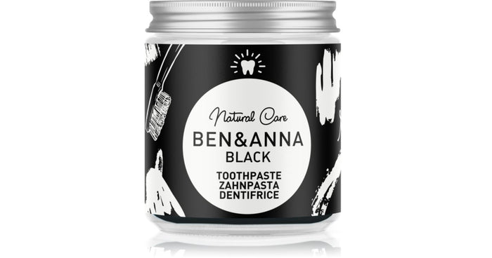 A jar of Ben & Anna Black Natural Care Toothpaste on a white background with a subtle reflection. The jar has a black label with white illustrations and a silver metal cap.