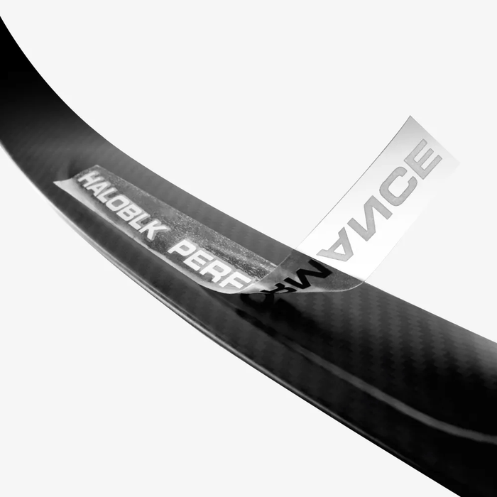 A close-up of the glossy carbon fiber surface of the HALOBLK Store Carbon Fiber Rear Spoiler for Tesla Model 3 shows a transparent sticker being peeled off, revealing the word PERFORMANCE and part of the HALOBLK brand name.