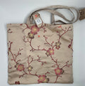 A beige foldable shopping tote bag with a floral pattern in shades of pink and red, featuring brown handles.
