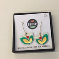 A pair of earrings designed with geometric shapes in white, turquoise, red, and bronze colors, presented on a black display card with the brand name 'DANA' at the top.