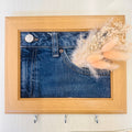 A square wooden frame hanging on a light wall, featuring a printed or mounted blue denim fabric panel with a pocket design, decorated with dried beige grasses, and equipped with three metal hooks below the frame.