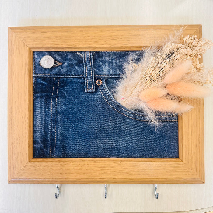A square wooden frame hanging on a light wall, featuring a printed or mounted blue denim fabric panel with a pocket design, decorated with dried beige grasses, and equipped with three metal hooks below the frame.
