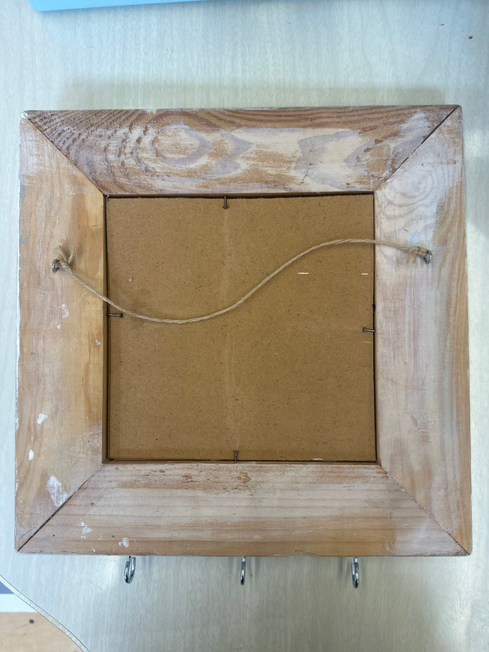 The back of a square wooden picture frame, showing the natural wood grain border, a brown backing board, and a hanging wire with metal hooks on the right side.