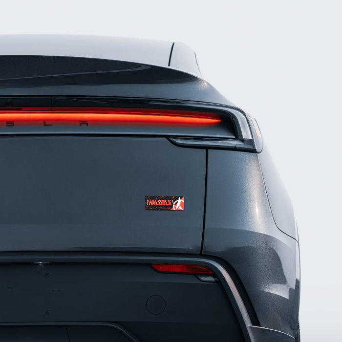 Close-up of the rear of a dark gray Tesla featuring the tail light, Tesla logo, and a HALOBLK Carbon Fiber Emblem from the HALO EFFECT Series by HALOBLK Store on the trunk.
