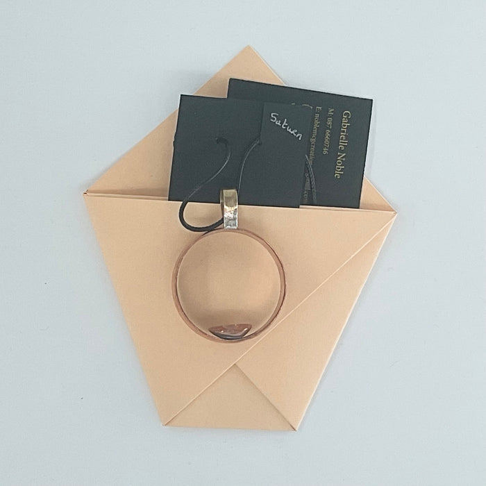 A rose gold ring with a silver-toned bail and black hangtag, displayed inside a beige paper envelope on a light gray background. The tag reads 'Sublime' and 'Catherine Nail'.