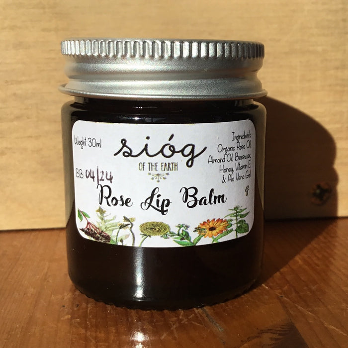 A jar of Sióg of the Earth Rose Lip Balm with a label showing the brand name, product name, and ingredients such as rose oil, almond oil, beeswax, honey, vitamin E, and aloe vera gel.