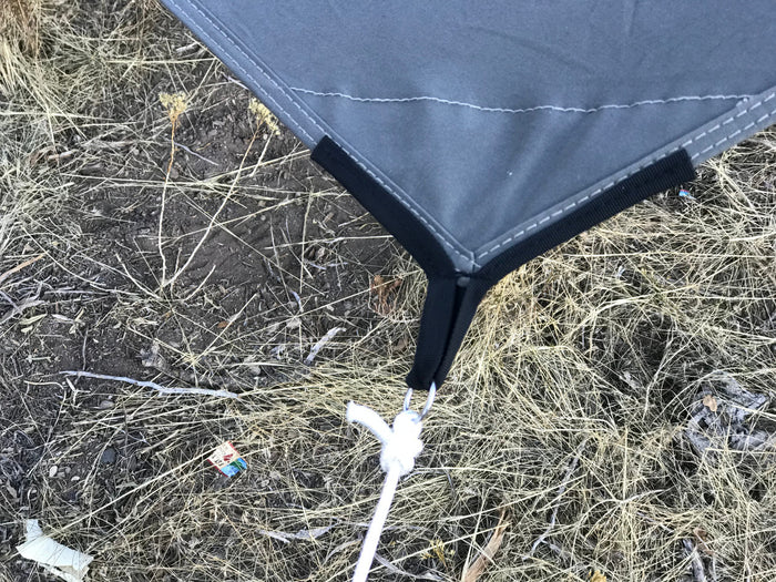 Close-up of a gray camping tarp corner with black elastic and a white tie-down in a natural setting.
