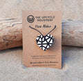 A necklace with a heart-shaped pendant featuring a black and white geometric pattern, displayed on a wooden surface with a brand tag attached.
