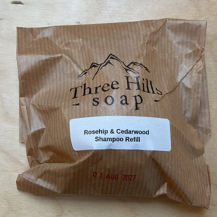A brown kraft paper refill pouch for Three Hills Soap, labeled 'Rosehip & Cedarwood Shampoo Refill', placed on a light wood grain surface.